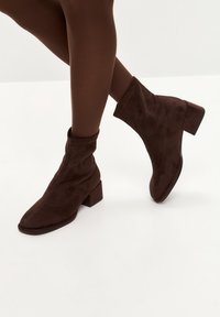 Bocanci scurți din suede maro, cu toc bloc, având un design lejer, vârf rotunjit și cusături tonale.