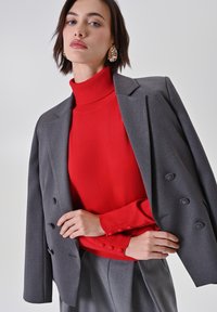 Grau gestreifter Doppelreiher-Blazer über einem roten Rollkragenpullover, mit glatter Textur und Knopfdetails. Das Model posiert vor einem neutralen Hintergrund.