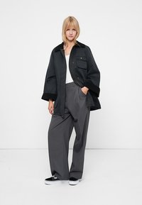 Jachetă neagră supradimensionată, cu două buzunare frontale, purtată peste un top alb, asociată cu pantaloni gri largi și adidași negri. Design simplu, detalii minime.