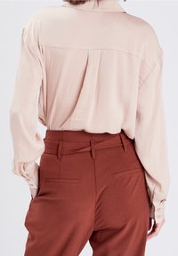 Blouse rose clair en soie avec une empiècement au dos, manches longues et poignets boutonnés, associée à un pantalon taille haute couleur rouille avec poches latérales.