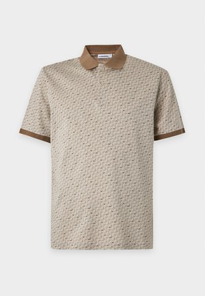 Homme debout portant un polo beige à motif, un pantalon large marron foncé et des baskets blanches sur un fond clair uni.