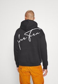 Sudadera negra con capucha y texto en cursiva blanca "Tu turno" en la parte posterior. Presenta un corte relajado y puños acanalados. Combinada con pantalones de pana naranja.