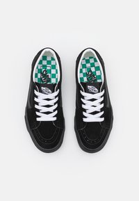 Pár černých tenisek Vans s bílými tkaničkami a zeleno-bílými kostkovanými stélkami, viděno shora na bílém pozadí.