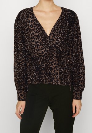 Kvinde iført en langærmet, omslagsbluse med brun og sort leopardprint og sorte bukser, stående mod en ensfarvet baggrund.