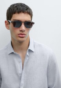 Sunglasses with dark lenses and a rectangular frame, featuring a brown and black color scheme. Worn with a light gray pinstriped shirt.