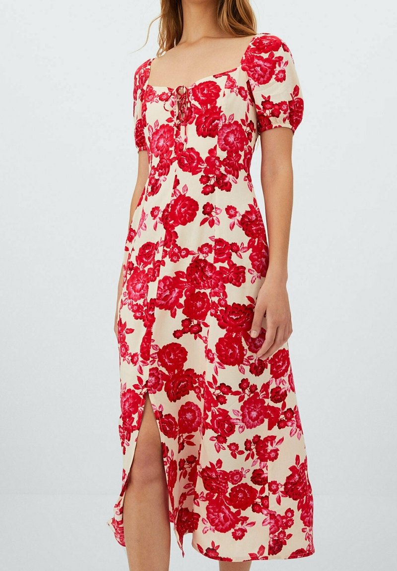 Femme portant une robe midi crème avec motif floral rouge, manches bouffantes, encolure carrée et fente sur le devant.