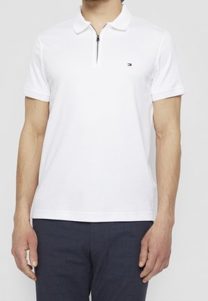 Mężczyzna ubrany w białą koszulkę polo z krótkim rękawem, z przednim suwakiem i małym logo na piersi, w połączeniu z ciemnymi, kraciastymi spodniami.