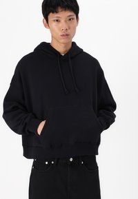 Hoodie cropped preto feito de tecido macio, com um bolso frontal, capuz com cordão e um corte descontraído. Usado com jeans pretos.