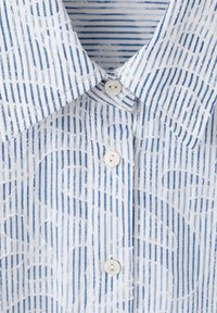 Gros plan d'une chemise blanche avec des rayures verticales bleues et un motif festonné blanc, avec un col et trois boutons blancs visibles.