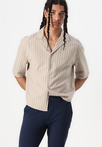 Chemise beige à manches courtes avec des rayures verticales bleu marine, dotée d'un col en pointe, d'une fermeture à boutons et d'un tissu texturé. Associée à un pantalon bleu marine.