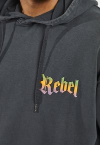 Sort hættetrøje med en farverig "Rebel" grafik i en fed, stiliseret skrift. Indeholder en hætte med snor og tekstureret stof.