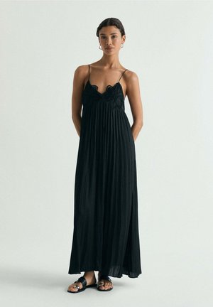 Mujer con un vestido largo negro plisado con tirantes finos y sandalias negras, de pie contra un fondo claro y sencillo.