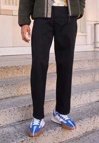 Czarne spodnie o prostych nogawkach z gładką fakturą, zestawione z niebiesko-białymi sneakersami z grubą podeszwą i kontrastującymi akcentami kolorystycznymi.