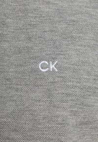 Tissu gris avec un tissage texturé. Présente un petit logo brodé "CK" en blanc. Vue en gros plan, mettant en évidence le matériau et le détail du logo.