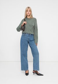 Pull vert olive avec broderies florales, associé à un pantalon en jean bleu à jambes larges et des escarpins à talons aiguilles noirs ; tenant un petit sac à main marron.