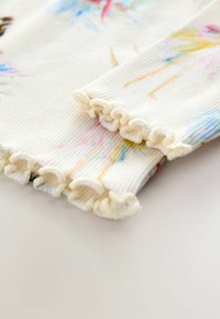 Randig vit textil med färgglada, akvarell-liknande mönster. De volangiga kanterna på ärmarna ger textur och detaljer till den övergripande designen.