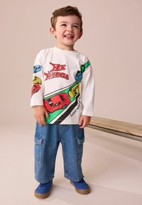 Biela dlhá košeľa s dlhým rukávom má grafiku "Hot Wheels" s farebnými autami, kombinovaná so svetlo modrými voľnými džínsami a modrými teniskami.