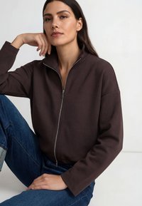 Rjava jakna na zadrgo z visokim ovratnikom, izdelana iz mehkega blaga. Ima sproščen kroj in dolge rokave, združena s svetlo modrimi kavbojkami.