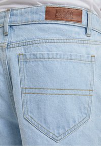 Világoskék farmernadrág, bőr márkajelzéssel, látható varrással és minimalista dizájnnal, két hátsó zsebbel.