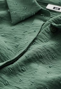 Gröntexturerad skjorta med mjukt, rynkat tyg, med krage och knapprad. Anmärkningsvärda upphöjda mönster genom hela materialet.