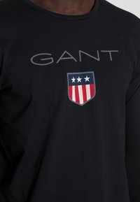 Schwarzes Langarmshirt mit dem Wort "GANT" in Grau und einem Schildlogo mit drei Sternen und roten Streifen in der Mitte.