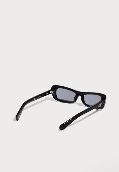 Lunettes de soleil rectangulaires noires avec des verres teintés foncés, posées sur une surface blanche, vues légèrement en angle arrière.