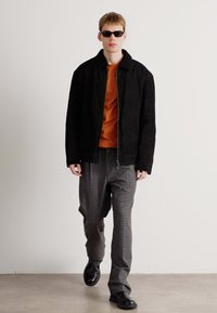 Giovane che cammina, indossando una giacca nera, una maglietta arancione, pantaloni grigi a righe, scarpe nere e occhiali da sole scuri su uno sfondo bianco semplice.