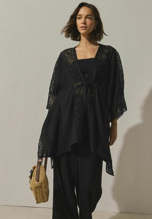 Tânără purtând un kimono negru din dantelă lejeră peste o ținută neagră, ținând o geantă țesută bej cu mărgele decorative, în picioare în fața unui perete simplu.