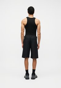 Άνδρας που στέκεται με την πλάτη γυρισμένη, φορά μαύρο αμάνικο πουκάμισο, μαύρα σορτς μέχρι το γόνατο, μαύρες κάλτσες και μαύρα αθλητικά σε λευκό φόντο.