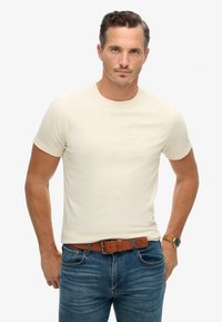 Camiseta de manga corta color crema hecha de tela suave; presenta un sutil logo "SuperDry" grabado en el pecho. Se lleva con jeans azules y un cinturón marrón.