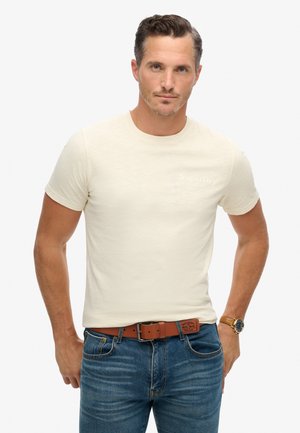 Crème-kleurig T-shirt met korte mouwen van zachte stof; heeft een subtiele geperste 'SuperDry' logo op de borst. Draag het met blauwe spijkerbroek en een bruine riem.