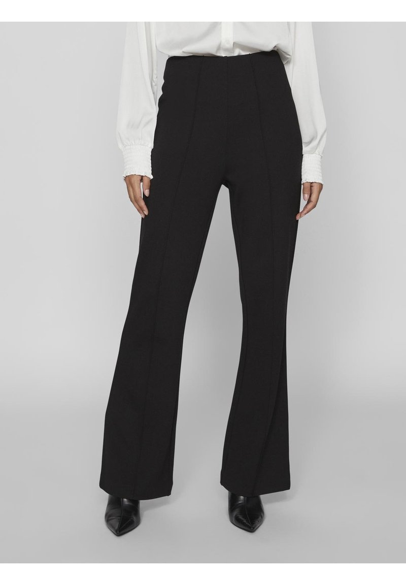 Svarta högmidjade byxor med vid benform, med vertikala sömmar och en slät textur. Kombination med en vit blus och svarta klackar.