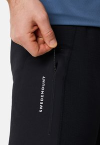 Schwarze Sporthosen mit einer seitlichen Reißverschlusstasche, versehen mit dem weißen "SWEDEMOUNT"-Logo. Das Material scheint leicht und dehnbar zu sein.