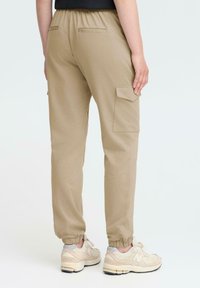 Cargo-bukser i lys beige stof med elastiske manchetter, der har sidelommer og en glat tekstur. Båret med lyse sneakers.