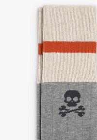Calcetines hasta la rodilla con parte superior beige y una franja naranja, y una parte inferior gris con un diseño de calavera y huesos cruzados en negro.