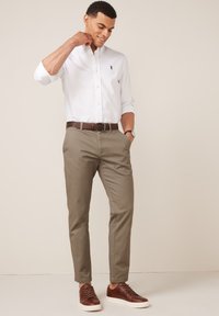 Witte button-up shirt met een klein logo, tan kleurige, getailleerde broek en bruine leren sneakers. De outfit heeft een bruine riem en horloge.