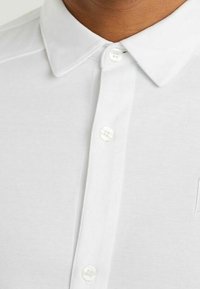 Biała koszulka polo z wyraźnym kołnierzykiem, gładką teksturą i subtelnymi przeszyciami. Zapinana na okrągłe, białe guziki.