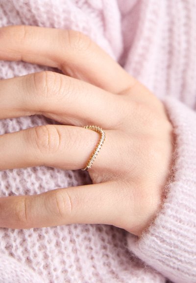 Anillo de oro con un diseño en zigzag, adornado con pequeñas piedras blancas, lucido en una mano de piel clara, sobre un suéter texturizado de color morado claro.