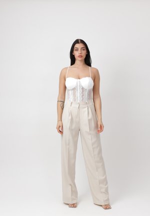 Donna con capelli lunghi e scuri che indossa un bustier bianco di pizzo e pantaloni larghi a vita alta color crema, in piedi davanti a uno sfondo chiaro e semplice.