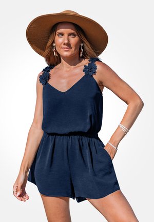 Femme portant une combinaison sans manches bleu marine avec des détails floraux sur les épaules, un chapeau marron à large bord et des bracelets blancs, posant avec la main dans la poche.