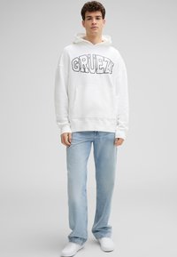 Witte hoodie met de tekst "GRÜEZI" in het zwart, voorzak, gecombineerd met lichtblauwe jeans en witte schoenen, staand tegen een grijze achtergrond.