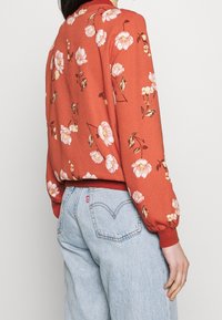 Veste bomber fleurie en orange brûlé avec poignets et ourlet élastiqués. Présente de grandes fleurs roses et crème avec des accents vert foncé.