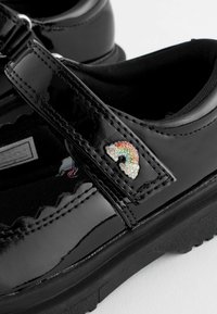 Chaussures vernies noires avec une sangle velcro, présentant un petit motif arc-en-ciel en cristaux sur le côté et un tissu noir texturé à l'intérieur.