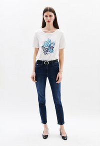 Witte T-shirt met korte mouwen en een blauwe bloemenprint, gecombineerd met donkerblauwe skinny jeans en zwarte hoge hakken.
