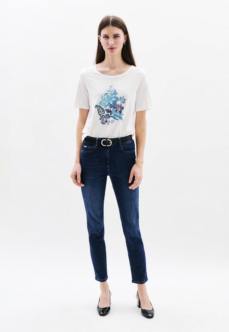Witte T-shirt met korte mouwen en een blauwe bloemenprint, gecombineerd met donkerblauwe skinny jeans en zwarte hoge hakken.