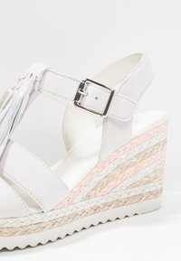 Sandale compensée blanche avec une plateforme texturée ornée de tresses beiges, blanches et roses. Une bride de cheville ajustable et des franges décoratives.