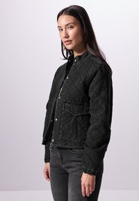 Chaqueta de denim negra acolchada con cremallera frontal, botones a presión y bolsillos laterales. Presenta un cuello alto y puños elásticos para un ajuste ceñido.