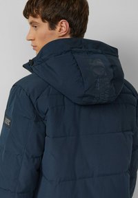 Granatowa kurtka puchowa z wysokim kołnierzem, odpinanym kapturem, przeszywanymi sekcjami oraz logo na ramieniu i plecach.