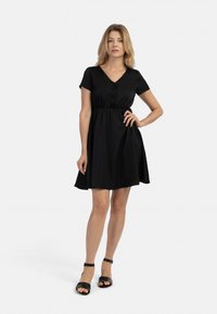 Rochie scurtă neagră cu mâneci scurte, decolteu în V, detalii cu butoane și talie elastică, fustă în formă de A; asortată cu sandale negre cu curea la gleznă.