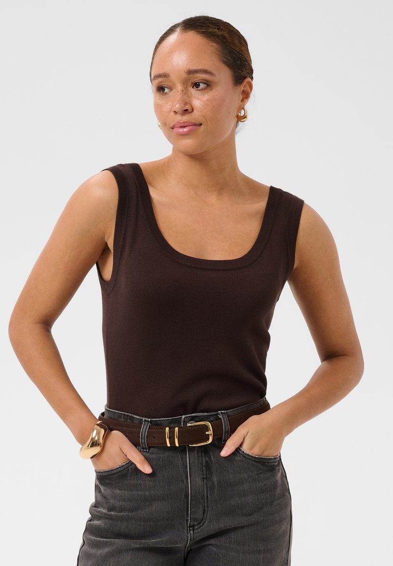 Femme à la peau brun clair et tachetée, portant un haut sombre sans manches, un jean noir, une ceinture marron, des boucles d'oreilles créoles en or et un bracelet en or, les mains dans les poches.